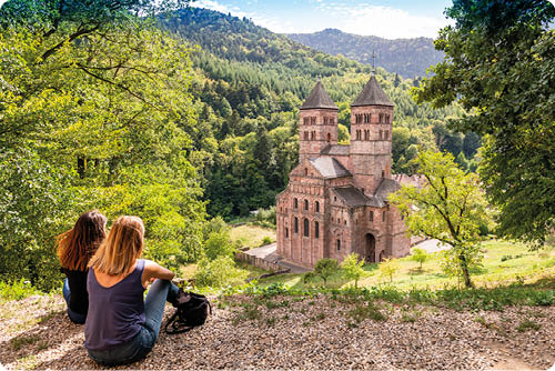 Zwei Personen sitzen auf einem Waldweg und blicken auf eine historische Kirche mit zwei T rmen, die von gr nen H geln und B umen umgeben ist. Beschreibung von KI generiert