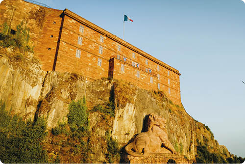Eine gro e steinerne L wenstatue sitzt vor einer Felswand, auf der ein historisches Geb ude mit einer wehenden Flagge steht. Beschreibung von KI generiert