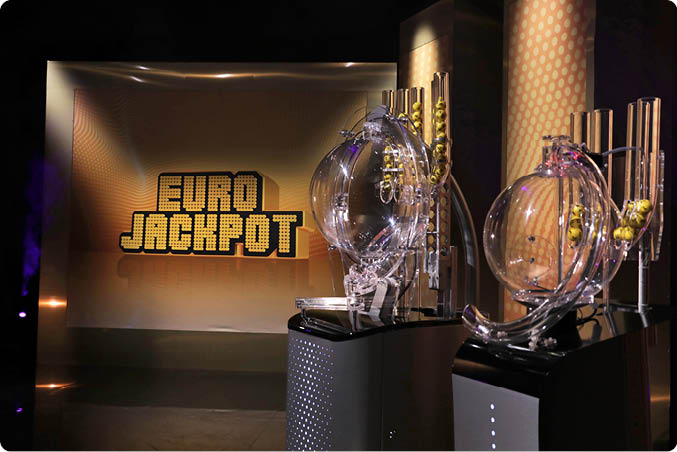 In einem Studio sind zwei transparente Lostrommeln vor einer beleuchteten Wand mit der Aufschrift Euro Jackpot zu sehen. Beschreibung von KI generiert