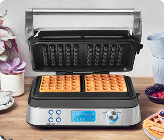 Ein moderner Waffeleisen mit zwei goldbraunen Waffeln steht ge ffnet auf einer Holzarbeitsplatte neben einer Pflanze, einer Tasse und einem Teller mit Beeren. Beschreibung von KI generiert